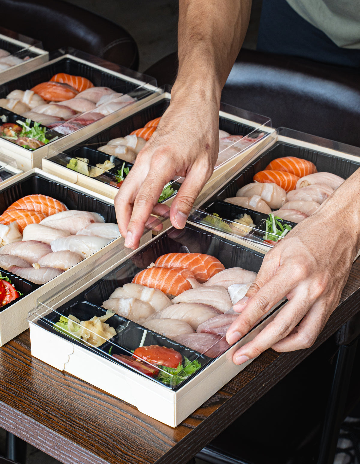 A close-up of a person's hands opening a clear-lidded wooden box filled with assorted sushi, including salmon and white fish pieces, set on a dark wooden table.